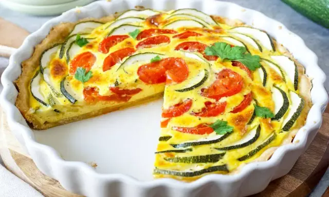 Herzhafte Quiche mit Zucchini und Tomaten, garniert mit Petersilie, in einer weißen Quicheform. Ein Stück fehlt.
