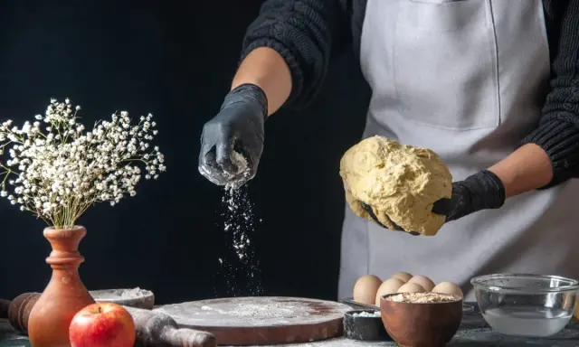 Hände in schwarzen Handschuhen streuen selbsttreibendes Mehl über Teig. Ein Apfel und Blumen sind auf dem Tisch.