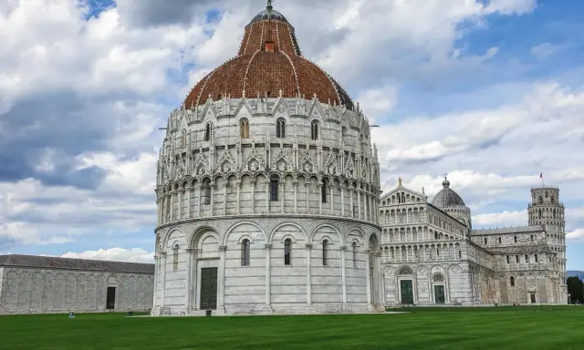Il Battistero, il Duomo e la Torre Pendente: ecco cosa vedere a Pisa in un giorno, un'iconica piazza con un prato verde e cielo nuvoloso.