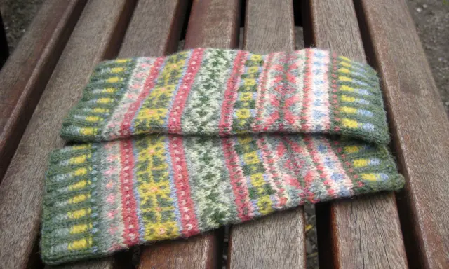 Två handledsvärmare i fair isle stickning med mönster i grönt, rosa, gult och blått ligger på en träbänk.