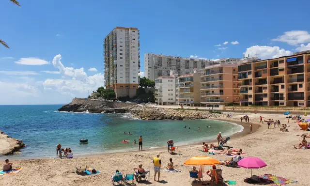 Sonniger Cullera Strand mit vielen Menschen, die sich im Meer und am Sand entspannen, im Hintergrund hohe Gebäude.