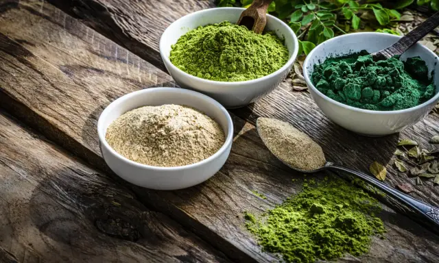 Drei Schalen mit Pulver auf Holz: hellbraun, leuchtend grün und dunkelgrün. Spirulina gesund und voller Nährstoffe.