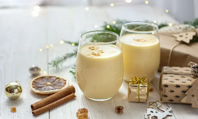 Zwei Gläser cremiger Cocktail mit Eigelb, garniert mit Zimt, umgeben von Weihnachtsdekorationen und Geschenken.