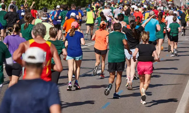 Tłum biegaczy pokonuje kolejne kilometry maratonu. Widać ich nogi w ruchu, kolorowe stroje i cienie na asfalcie.
