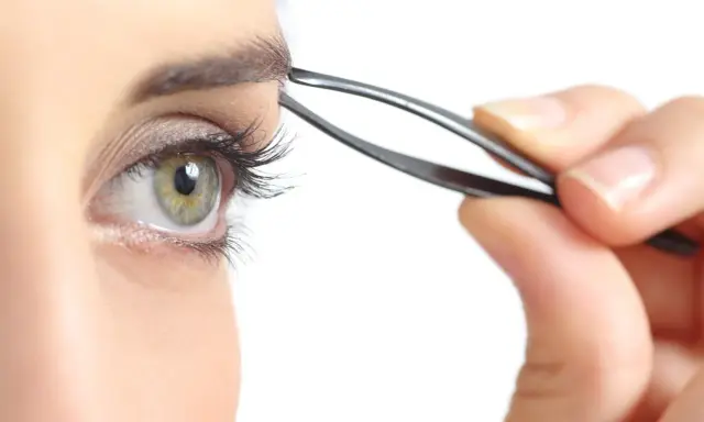 Une main utilise une pince à épiler pour parfaire l'épilation des sourcils d'un œil vert aux longs cils.