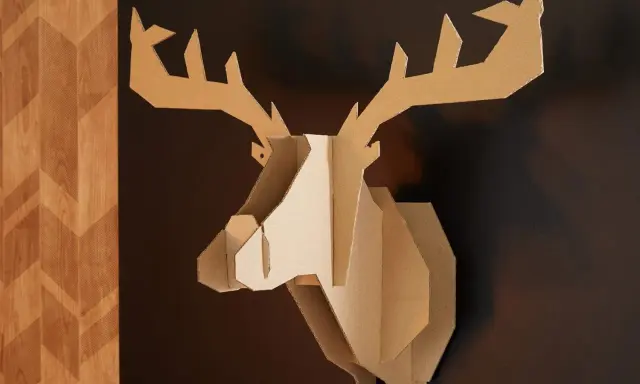 Trophée de cerf stylisé, œuvre de bricolage avec du carton de récupération, accroché sur un mur sombre à côté d'un panneau décoratif en bois.