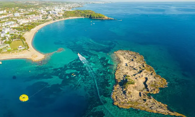 Widok z lotu ptaka na wybrzeże z hotelami, plażą i turkusową wodą. Widać łódź, parasailing i skaliste wysepki, które są świetnymi okurcalar atrakcje.