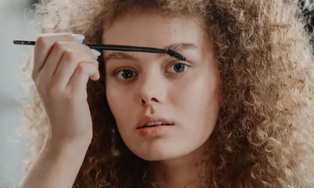 Une jeune femme aux cheveux bouclés applique un produit sur ses sourcils décolorés avec une brosse.