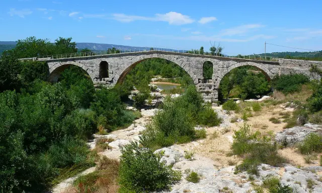 Vieux pont de pierre à arches sur une rivière asséchée, entouré de végétation luxuriante.