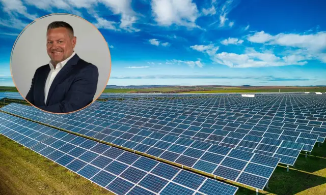 Uśmiechnięty mężczyzna w garniturze na tle farmy fotowoltaicznej. Obraz symbolizuje produkcję energii z fotowoltaiki w poszczególnych miesiącach.