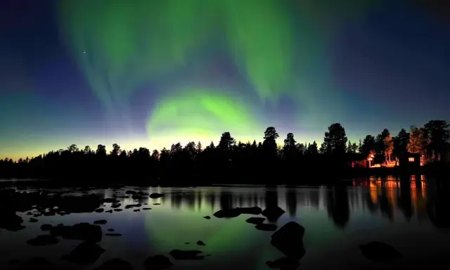 Norrsken dansar över en vinterlandskap vid Kiruna. En skogskant och en sjö med stenar speglar himlens magiska ljus.