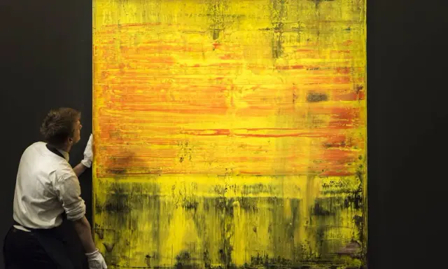 A man in white gloves carefully handles a large abstract painting with vibrant yellow and orange hues, a testament to Gerhard Richter's net worth.