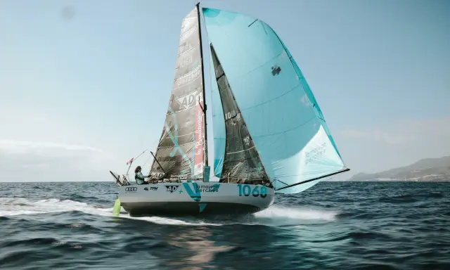 Un voilier de course, le numéro 1068, navigue sur une mer agitée. Les voiles, l'une noire et l'autre bleu clair, portent le nom "la maison perchée". Thais Le Cam est à la barre.