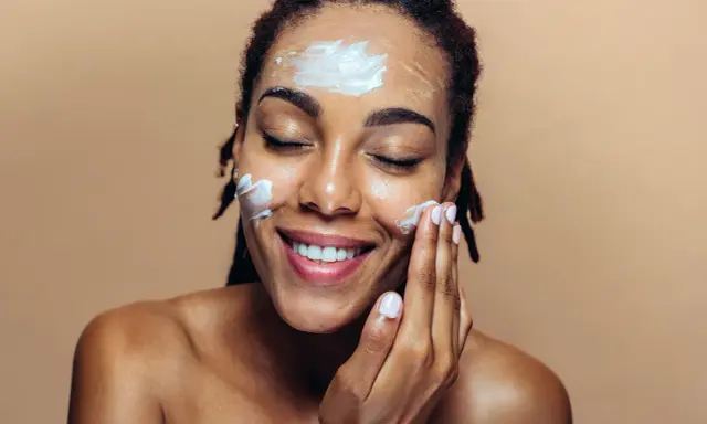 Une femme applique de la crème sur son visage. Elle sourit, les yeux fermés. Elle se demande que veut dire indice 50 pour sa protection solaire.
