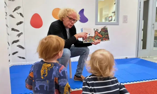 En man med lockigt hår läser en bok för två små barn. Bilden visar en humlan djojj på en morot i boken.