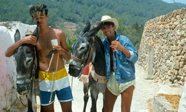 Tony Pike Ibiza: due ragazzi con asini, uno con pantaloncini colorati, l'altro con giacca di jeans e cappello di paglia, sorseggiano bevande.
