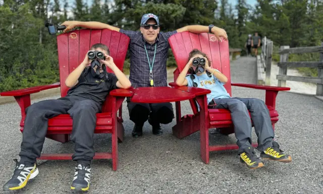 Un padre y sus dos hijos disfrutan de la naturaleza en Canadá con niños, observando el paisaje con binoculares.