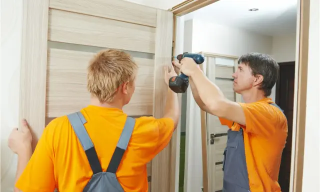 Dwóch mężczyzn montuje drzwi. Jeden przytrzymuje, drugi przykręca. Pomoc w sytuacji, jak naprawić spuchnięte drzwi.