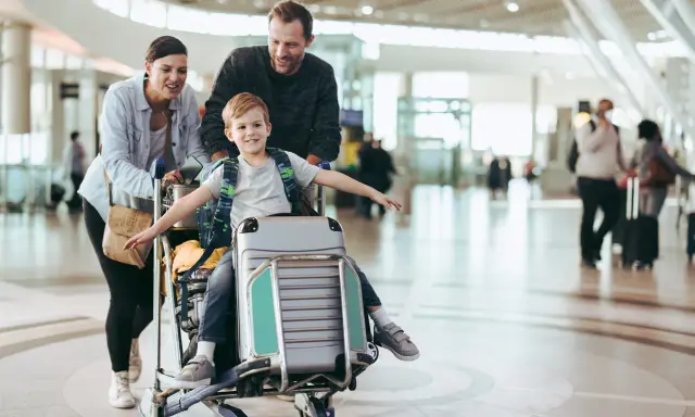 Familj på flygplatsen med ett barn sittandes på handbagage. De är glada och redo för sin resa.