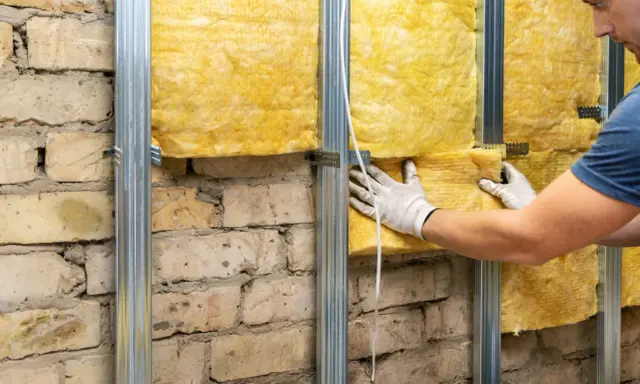Pose d'isolant jaune sur une structure métallique fixée à un mur de briques. L'isolation intérieure est en cours.