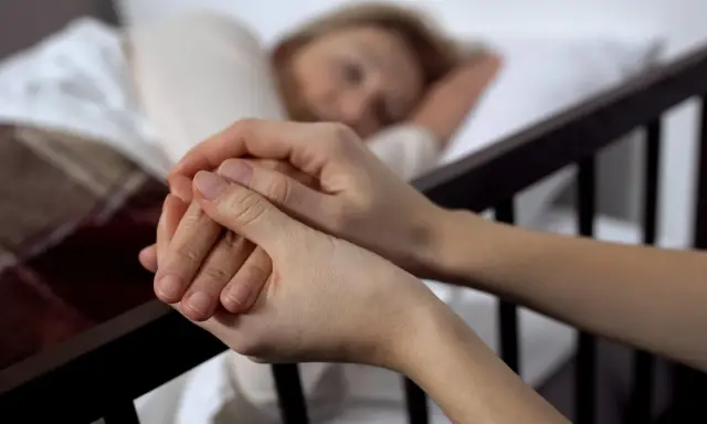 Dwie dłonie splatają się, jedna z nich spoczywa na ramieniu śpiącej osoby. To obraz towarzyszenia w trudnych chwilach, gdy rozważana jest eutanazja co to.