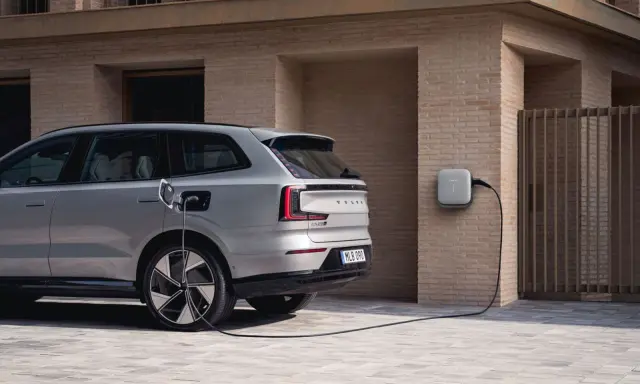 Srebrny, duży SUV Volvo EX90 podłączony do ładowarki ściennej. Nowoczesny design i ekologiczna energia.