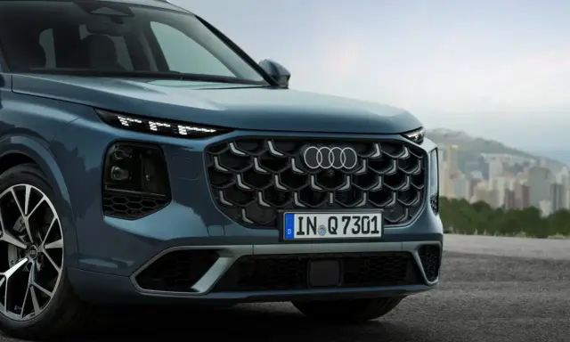 Przód eleganckiego Audi Q7 3.0 TDI w kolorze granatowym, z charakterystycznym grillem i światłami LED, na tle miejskiego krajobrazu.