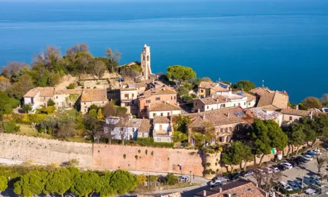 Borgo antico con campanile affacciato sul mare Adriatico. Il centro storico del lungomare Gabicce Mare, con tetti rossi e vegetazione rigogliosa, si estende verso l'azzurro intenso dell'acqua.