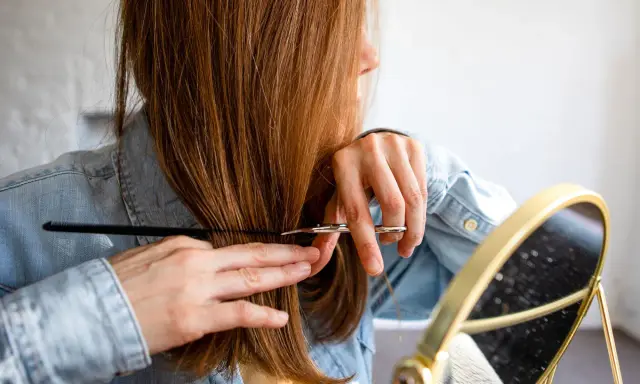 Frau mit langen, braunen Haaren, die sich im Spiegel die Haare schneidet.