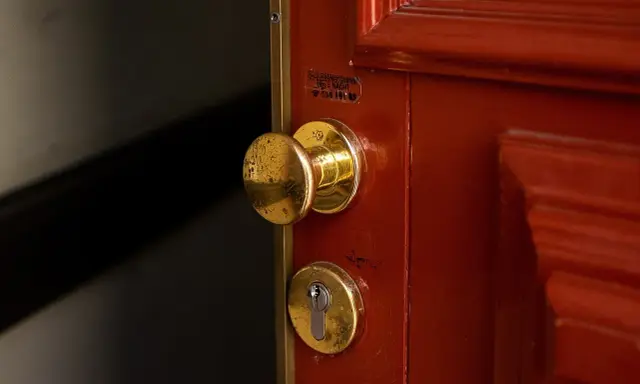 Poignée et serrure dorées sur une porte rouge. Une étiquette indique un service de serrurerie, utile en cas de porte forcée, pour l'assurance propriétaire ou locataire.