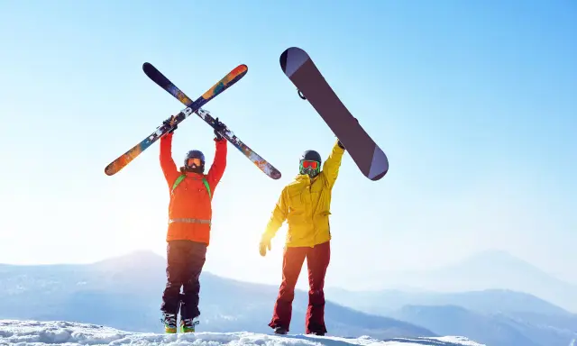 Dwóch narciarzy na szczycie góry, jeden z nartami, drugi z deską snowboardową, cieszy się słońcem i widokiem.