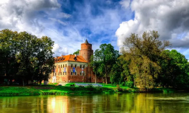 Malowniczy zamek w Łódzkiem nad rzeką, otoczony zielenią i chmurami.