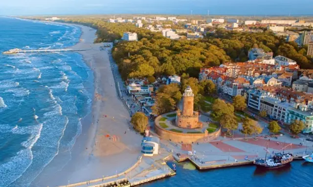 Malownicze miasta nad morzem z latarnią morską, plażą i molo. Widać port z żaglowcem i falujące morze.