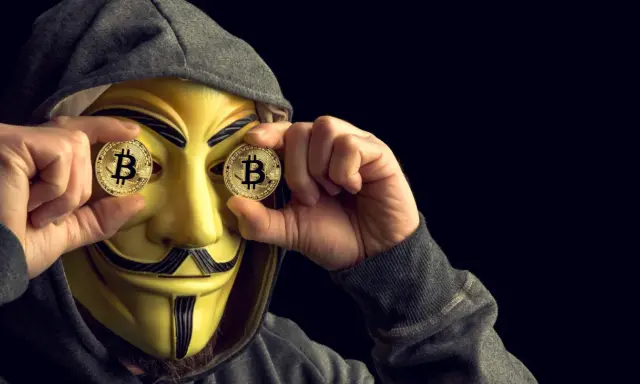 Osoba w masce Guya Fawkesa trzyma monety z Bitcoinem przed oczami. Czy MoonPay jest bezpieczny?