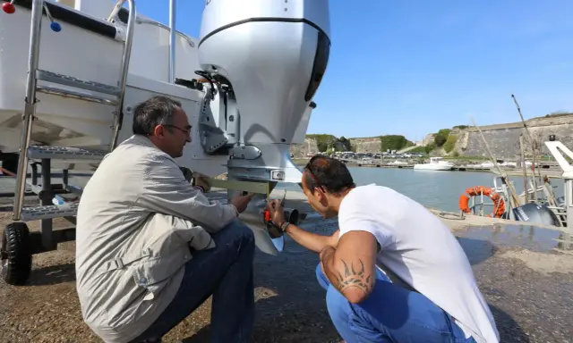 Deux hommes vérifient le montage moteur hors bord sur tableau arrière d'un bateau, près d'un port.