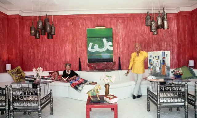 Interni di case anni '70: una donna legge sul divano bianco, un uomo mostra un quadro. Pareti rosse, lampade a grappolo.