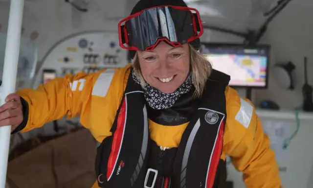 Alexia Barrier, souriante, porte une combinaison jaune et un gilet de sauvetage, prête pour une aventure maritime.