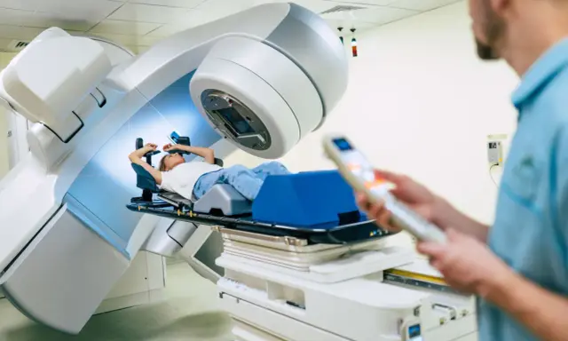 Ragazza sdraiata su lettino per radioterapia, tecnico in primo piano controlla il macchinario. Speranza di guarigione, ma con il timore dei radioterapia danni permanenti.