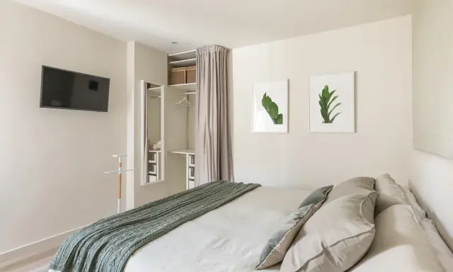 Ein offener Kleiderschrank, der wie ein Kleiderschrank selber bauen mit Vorhang wirkt, mit Regalen und Kleiderstange. Daneben ein Spiegel und ein Bett mit grüner Decke.