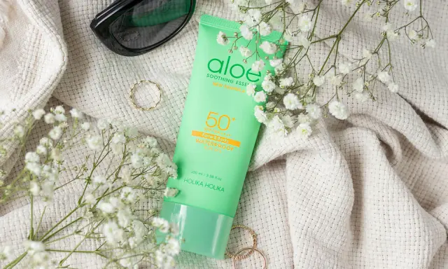 Zielona tubka z filtrem przeciwsłonecznym SPF 50 z aloesem, otoczona delikatnymi białymi kwiatami i okularami przeciwsłonecznymi.
