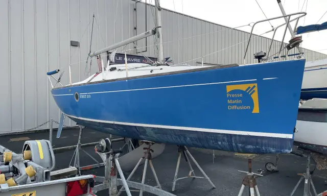 Voilier bleu FIRST 210 sur bers, prêt pour la saison. Le logo "Presse Matin Diffusion" est visible sur la coque.