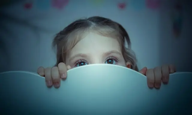 Occhi spalancati nel buio, un bambino si nasconde dietro il bordo del lettino, vittima di un pavor notturno.