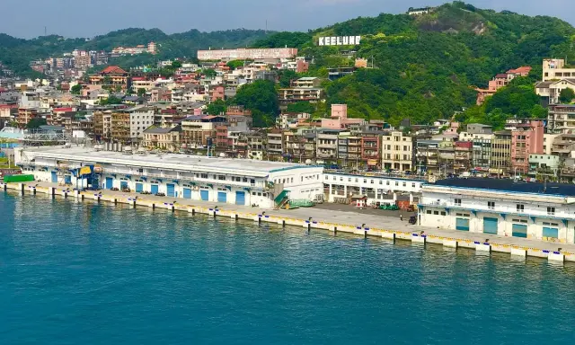Vista panoramica del porto di Taiwan, con edifici colorati che si arrampicano sulla collina verde. La scritta "KEELUNG" domina la cima.