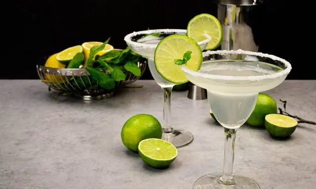 Zwei Margaritas mit Salzrand, garniert mit Limettenscheiben und Minze. Limetten und Minze in einer Schale im Hintergrund. Perfekt für ein Margarita Rezept.