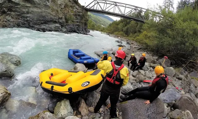 Grupa w kamizelkach ratunkowych i kaskach przygotowuje się do raftingu na rwących rzekach w Austrii. Dwie dmuchane łodzie czekają na brzegu.