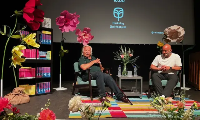 Jan Guillou delar fakta på Rinkeby Bokfestival. Scenen är dekorerad med stora blommor och bokhyllor.