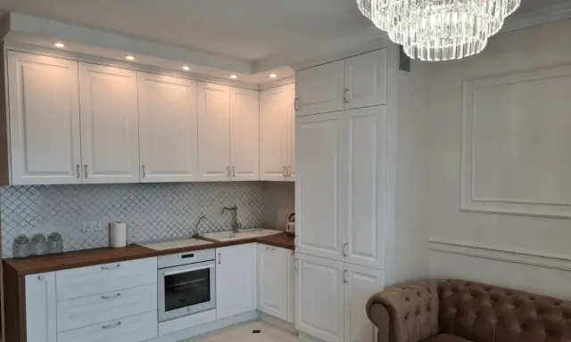 Elegancka kuchnia w stylu angielskim z białymi frontami, drewnianym blatem i kryształowym żyrandolem.