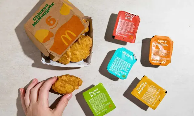 Ręka sięga po McNuggeta z pudełka. Obok sosy: Tangy BBQ, Spicy Buffalo, Ranch, Sweet 'N Sour, Honey Mustard. 20 McNuggets kcal to kusząca wizja.