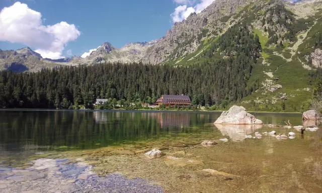 Spokojne Popradske Pleso odbija góry i las. Kamienie w płytkiej wodzie tworzą ścieżkę.