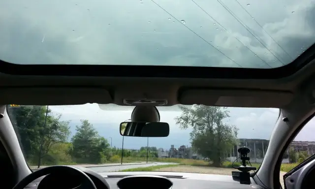 Widok przez panoramiczny dach samochodu. Krople deszczu na szybie, droga i drzewa w tle.